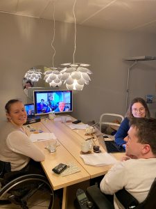 Regine, Mathias og Anna sitter rundt et bord med en stor skjerm, der resten av styret er med digitalt. 